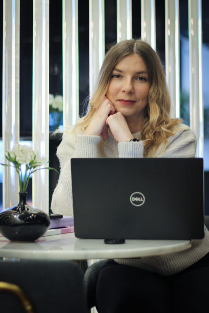 Pikkade blondide juustega naine istub laua taga, Dell sülearvuti ees, lõug käele toetatud - võib-olla plaanib personaaltreeneri virtuaalassistent. Tema kõrval on väike must vaas valgete lilledega ja taustal helendavad vertikaalsed tuled.