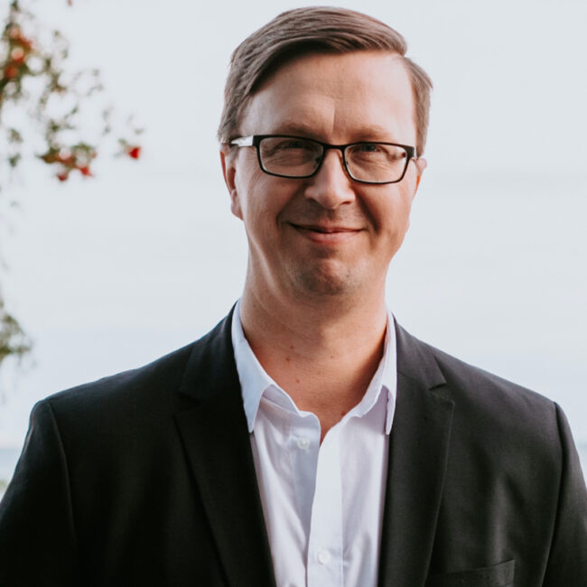 Mees, kes kannab prille ja tumedat ülikonnajakki üle valge särgi, seisab õues ja naeratab kaamerasse. Tema taga on ähmased taimed ja helge taust, jäädvustades inspiratsioonipäev 2026 vaimu.