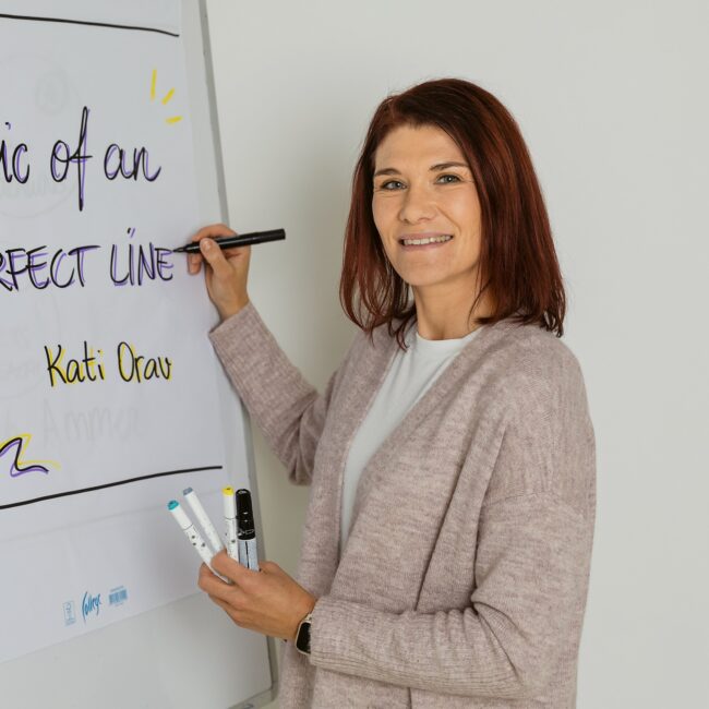 Inspiratsioonipäeval aastal 2026 seisab õlgadeni pruunide juustega naine flipchart'i juures, millel on kirjas "Ebatäiusliku joone maagia" ja "Kati Orav". Ta hoiab käes markerid ja naeratab kaamerasse.
