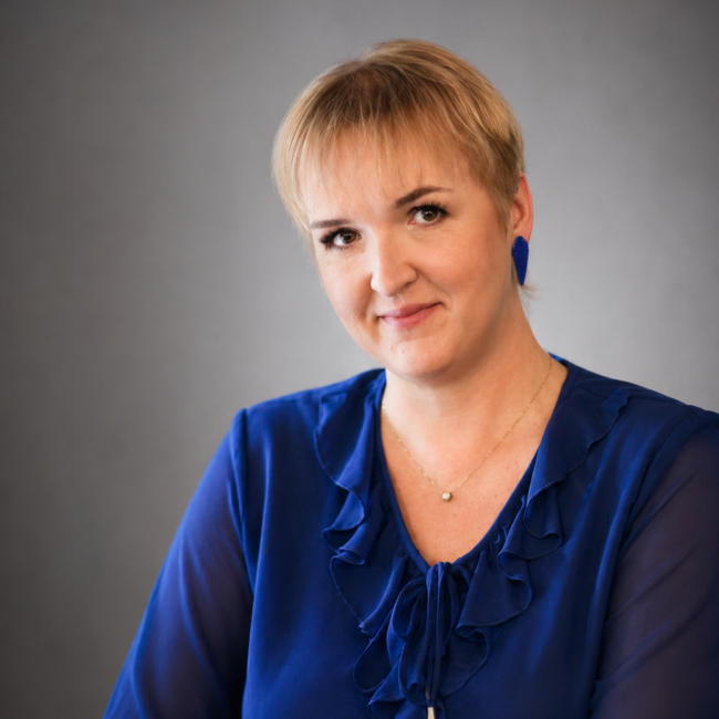 Lühikeste blondide juustega inimene kannab sinist rüüsidega pluusi, siniseid kõrvarõngaid ja kuldset kaelakeed, istub lihtsal halli taustal ja vaatab kaamerasse kerge naeratusega - ideaalne inspiratsioonipäev või Virtuaalassistent profiilile aastal 2026.